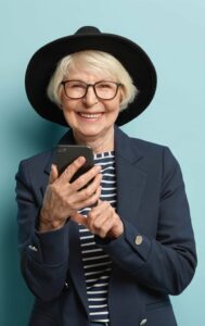 Cheerful female pensioner learns how to use smartphone, types notification, wears hat and jacket, connected to wireless internet. Charming granny stays in touch with gadget, isolated on blue wall